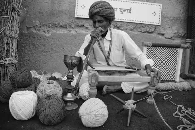 Black and white photo: an older man in a turban smokes a water pipe while weaving.